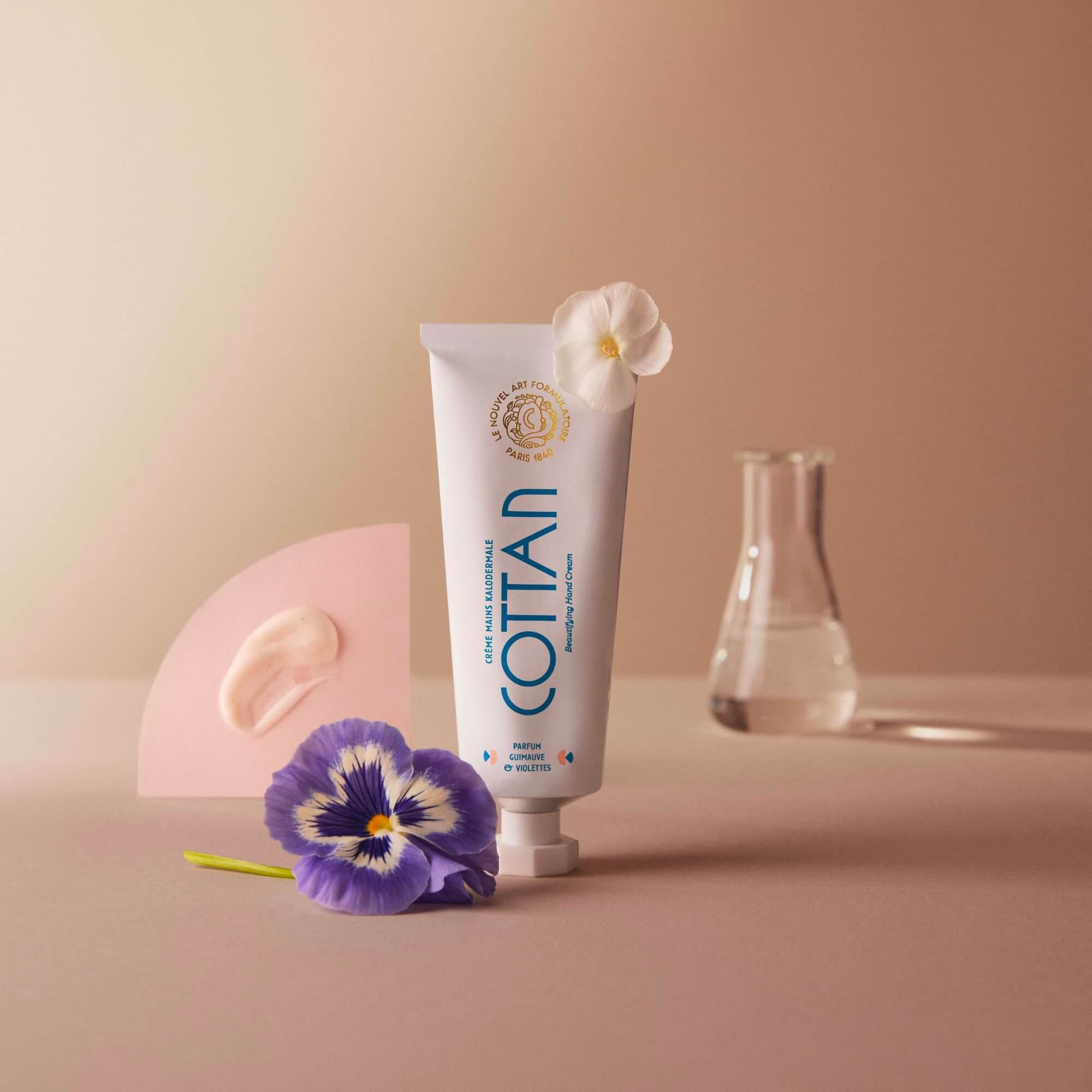 Tube de crème Kalodermale pour les mains COTTAN mis en scène avec une fleur de pensée, une goutte de crème sur un support rose, et des instruments de laboratoire en arrière-plan, sur un fond neutre.
