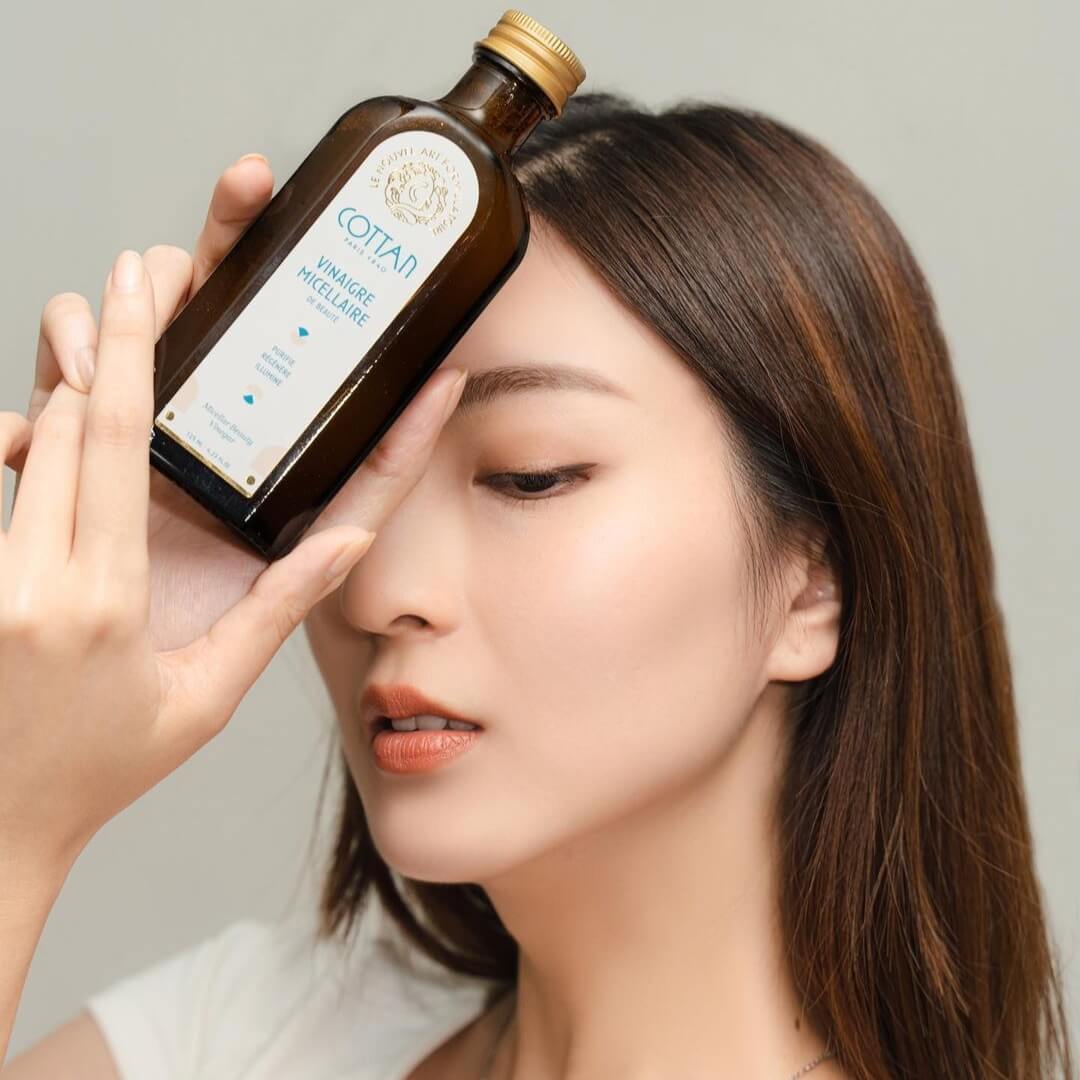 Portrait d’une femme tenant le vinaigre micellaire de beauté COTTAN contre son front, mettant en valeur le flacon ambré et l’étiquette iconique. Image lifestyle illustrant l’univers sensoriel et raffiné de COTTAN Paris 1840, associant pureté de la peau, douceur du teint et élégance naturelle.