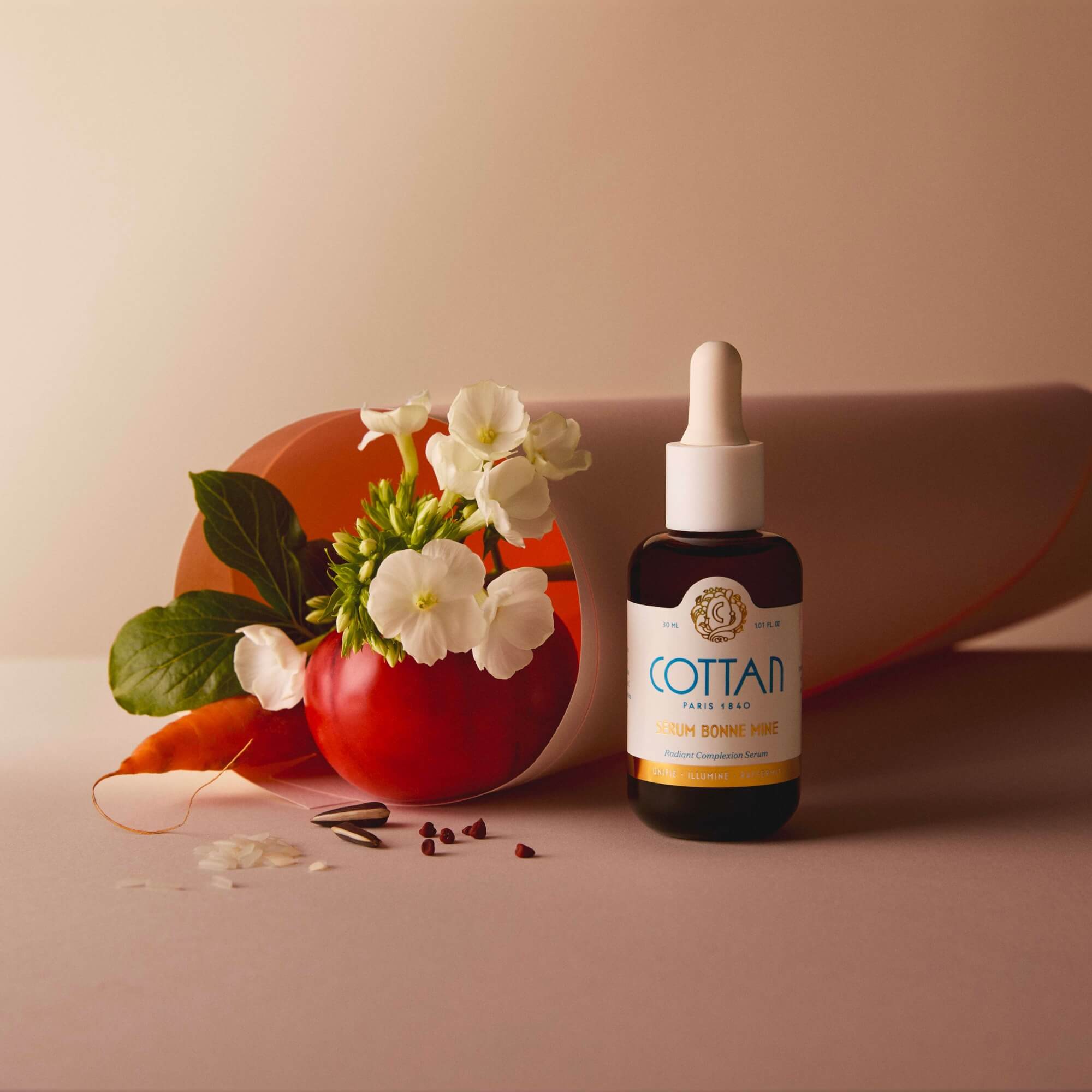 Flacon de sérum COTTAN "Bonne Mine" avec vitamine C placé devant une composition de tomate, carotte, graines et pétales, évoquant les ingrédients naturels du sérum, sur un fond neutre.