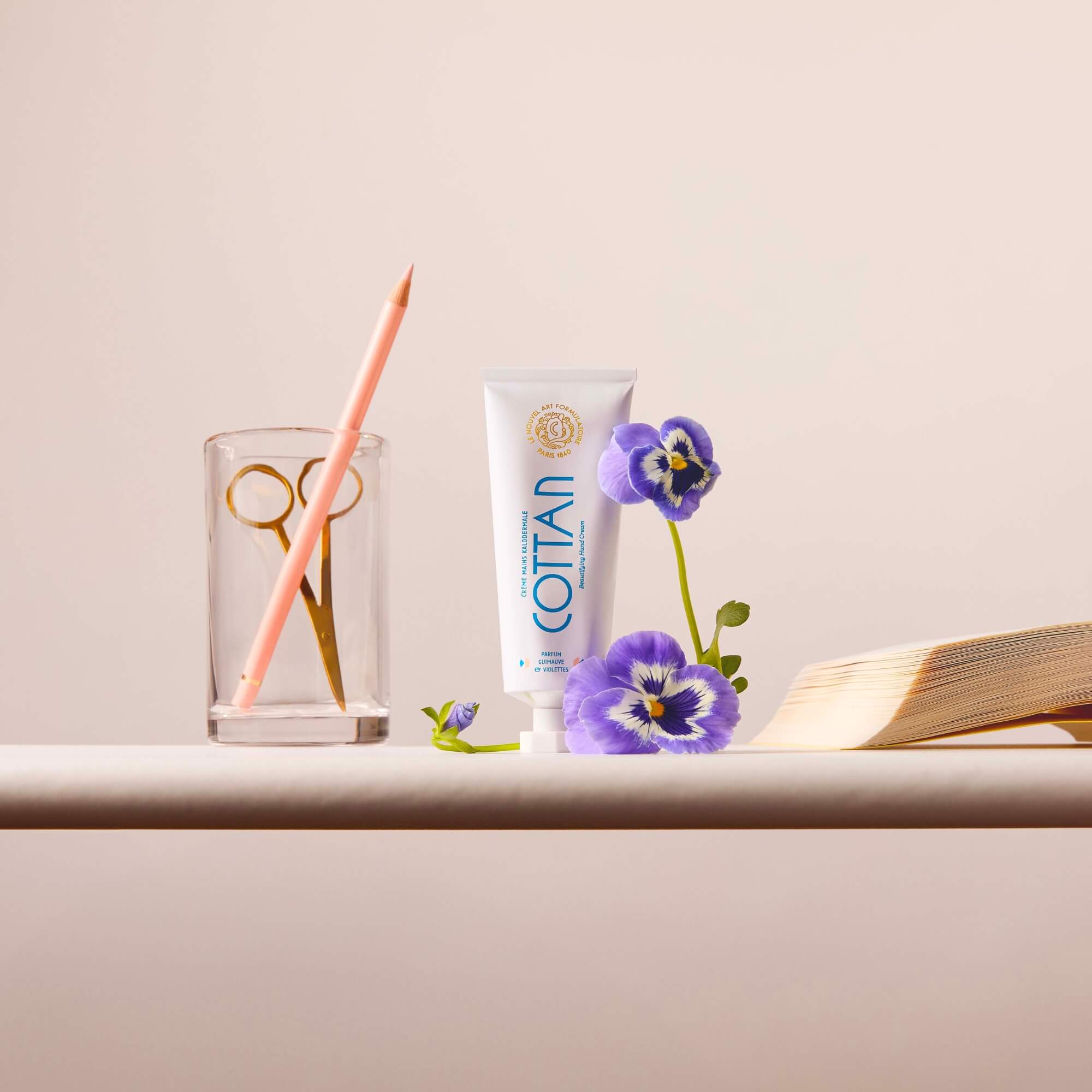 Tubo de crema Kalodermale para las manos COTTAN acompañado de flores de pensamiento, tijeras doradas y un lápiz en un vaso, presentado sobre un fondo neutro.