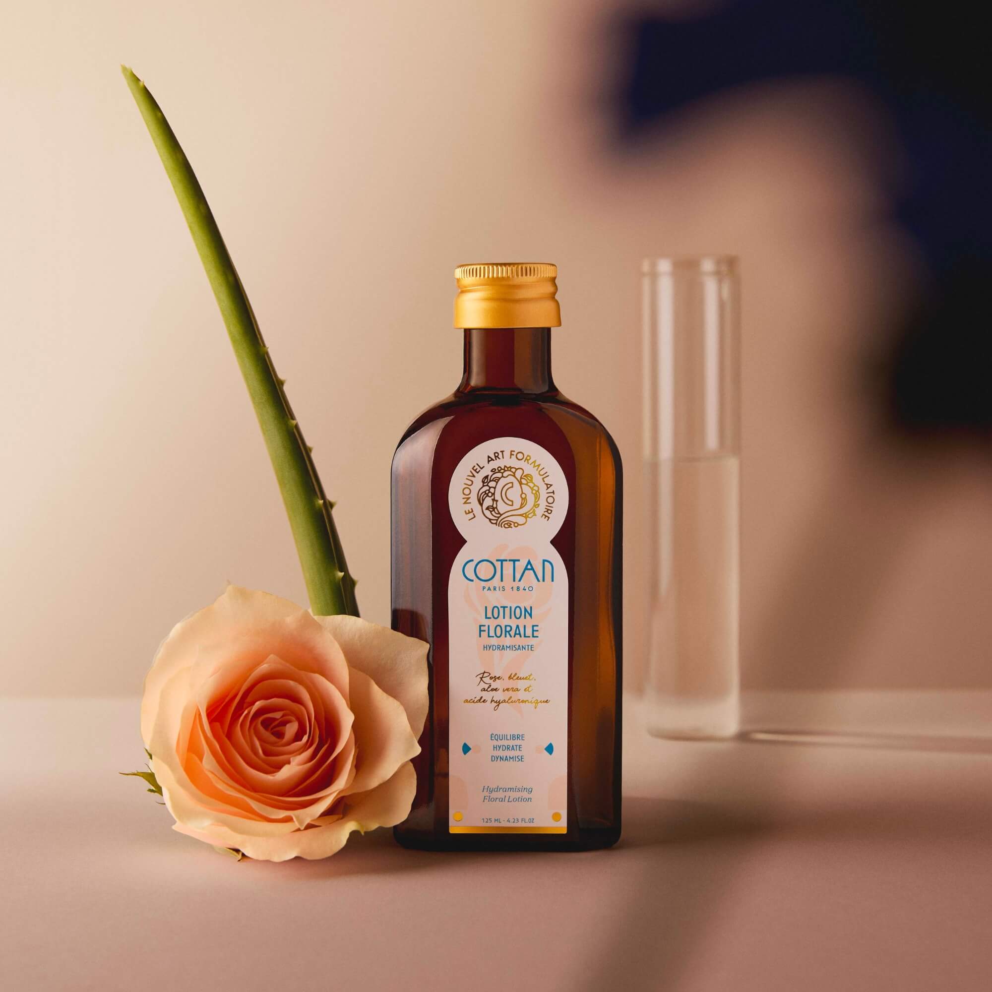 Botella de cristal de loción floral hidratante COTTAN colocada junto a una hoja de aloe y una rosa, sobre un fondo neutro con un tubo de ensayo al fondo.