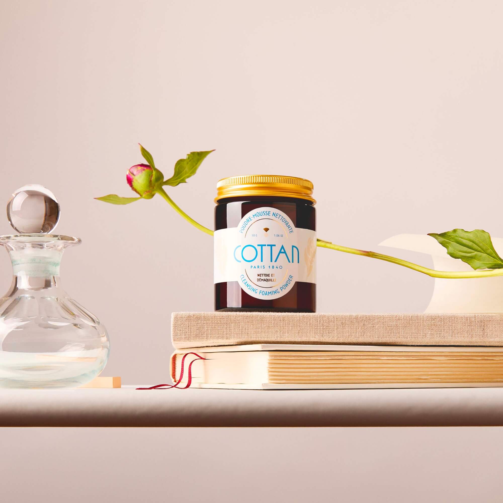 Glass jar of COTTAN cleansing powder placed on a book, with a rosebud next to it and a glass bottle in the background, on a neutral background.