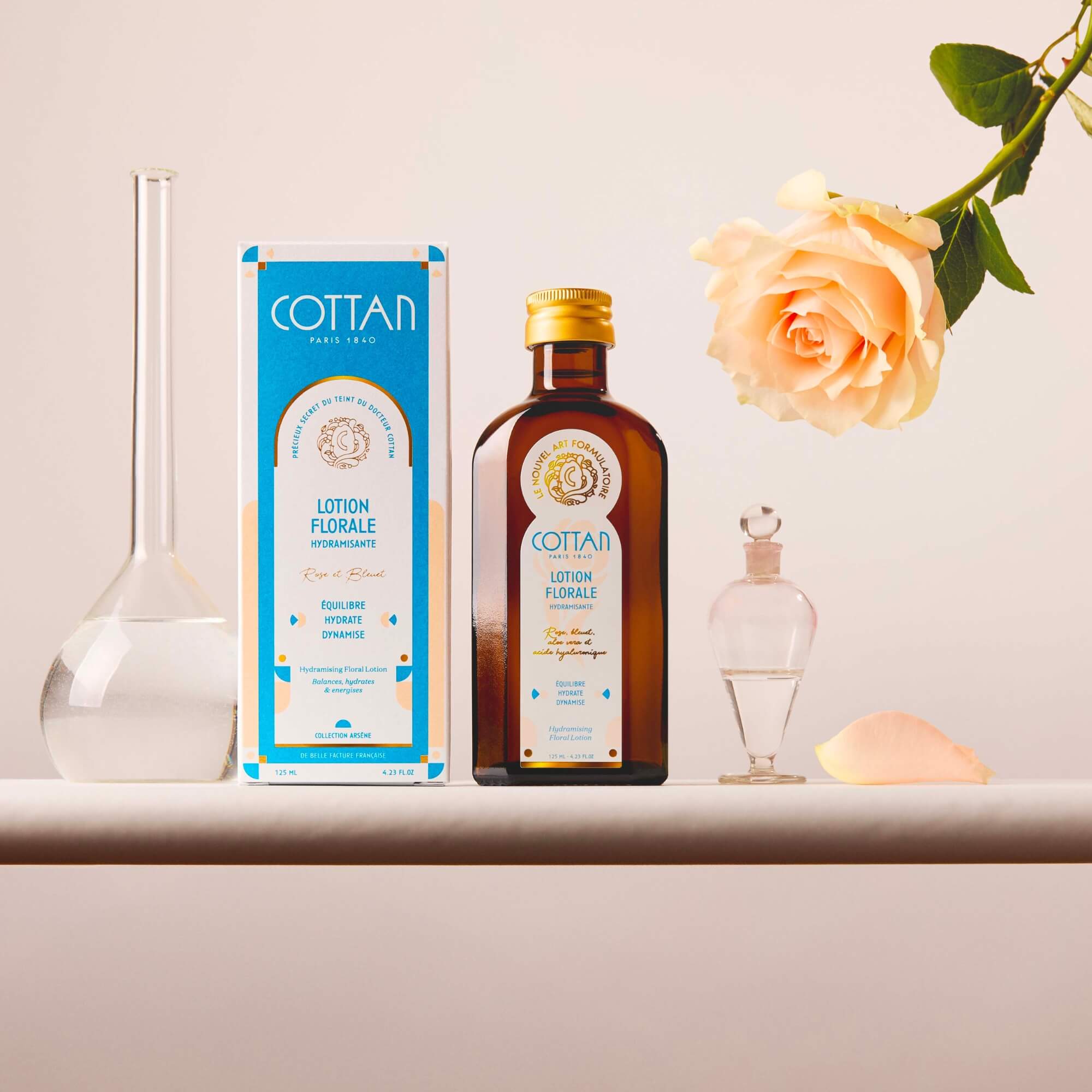 Botella de cristal de loción floral hidratante COTTAN junto a su caja de embalaje, flanqueada por un frasco de cristal y una rosa abierta sobre un fondo neutro.