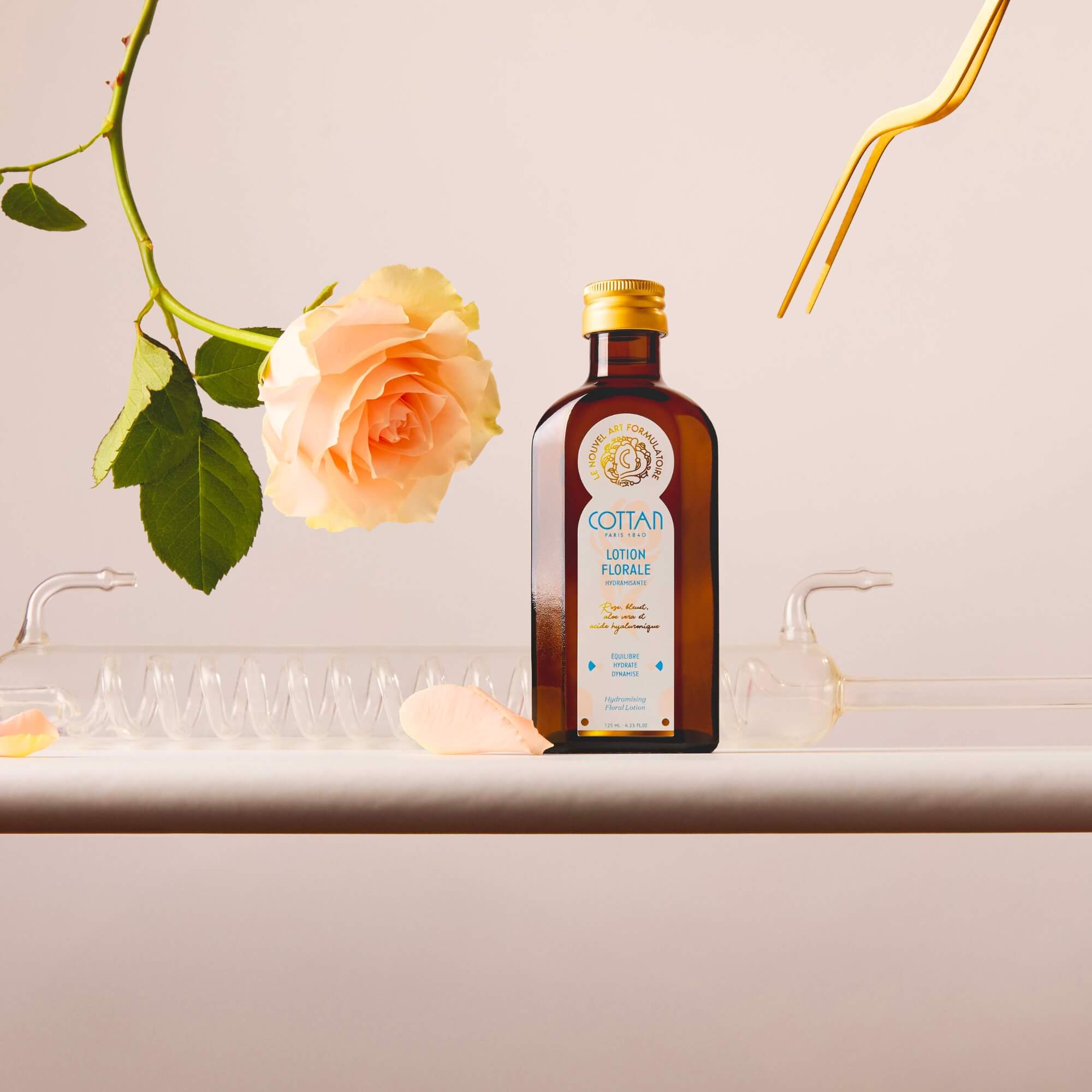 Botella de cristal de loción floral hidratante COTTAN con una rosa abierta suspendida sobre ella, acompañada de una pinza dorada, sobre un fondo neutro.