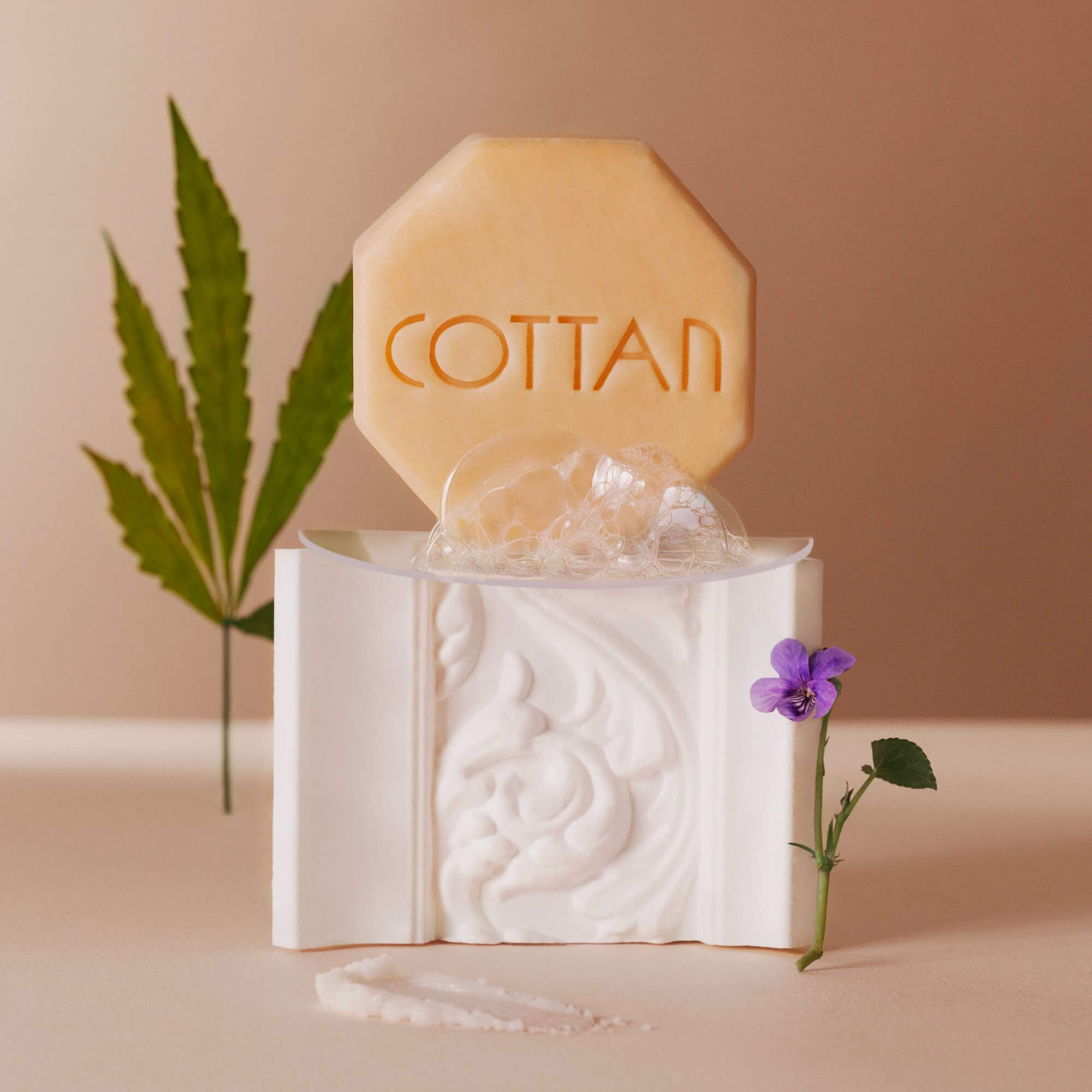 Composición artística en la que se muestra el jabón facial Dulcifié COTTAN Paris 1840, delicadamente colocado sobre un escultural estuche y acompañado de una espuma aérea. Los elementos vegetales y florales evocan la naturalidad y la suavidad de la fórmula, mientras que la puesta en escena depurada subraya la sensorialidad del gesto. Una imagen poética que traduce un ritual de limpieza relajante, que combina pureza, elegancia y herencia dermocosmética.