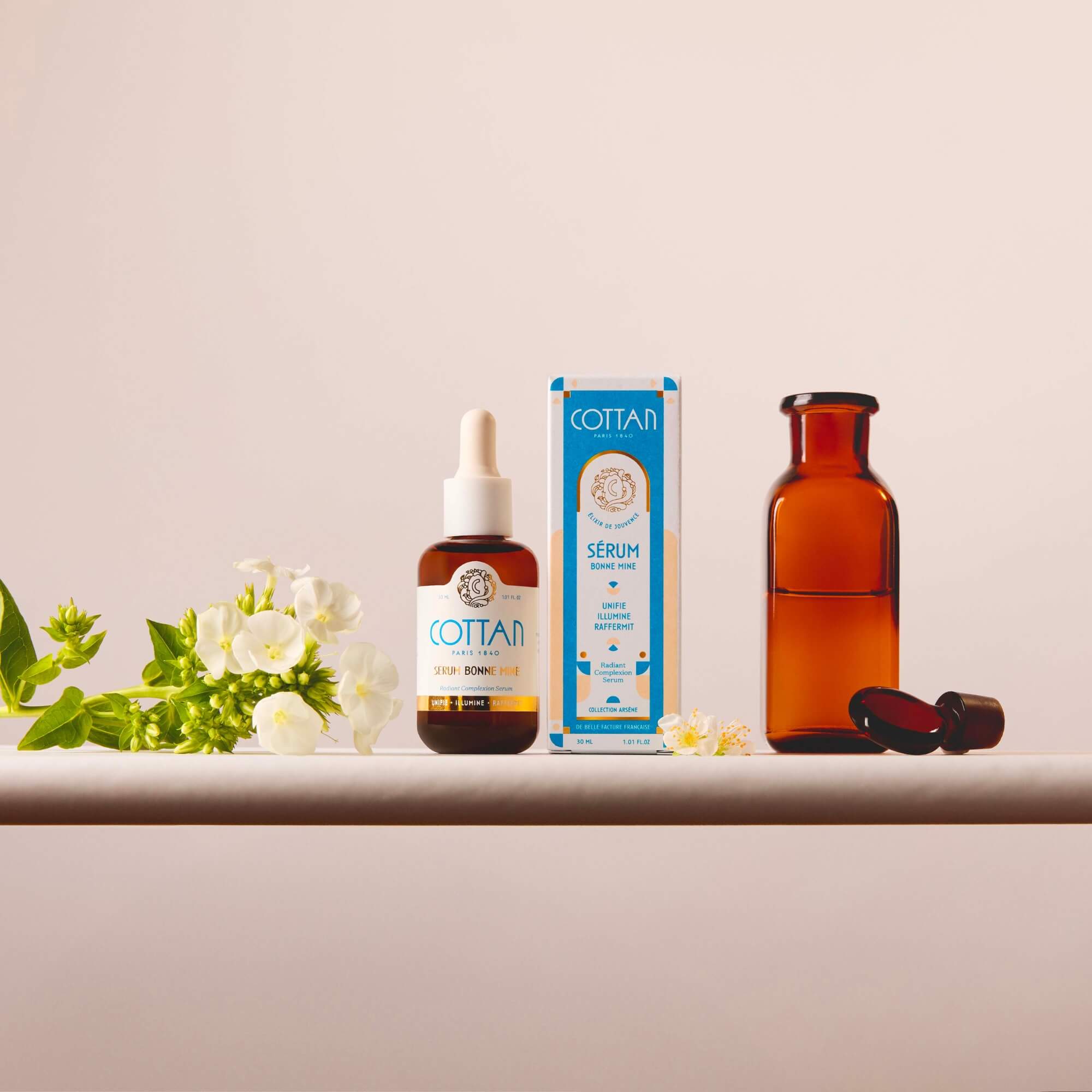 Frasco de sérum COTTAN «Bonne Mine» con vitamina C junto a su caja de embalaje, acompañado de una botella de cristal naranja y flores, sobre un fondo neutro.